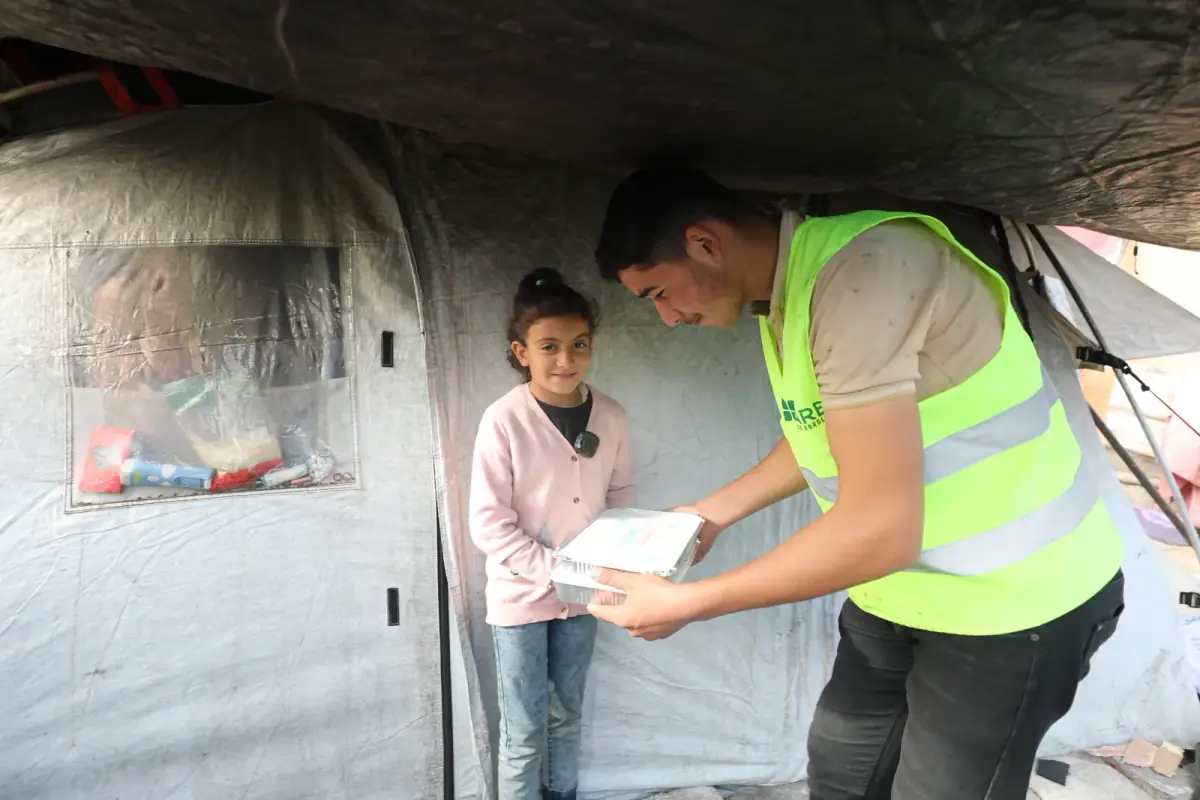 A CBB volunteer hands a hot-meal box to a young girl at her tent in a Gaza camp