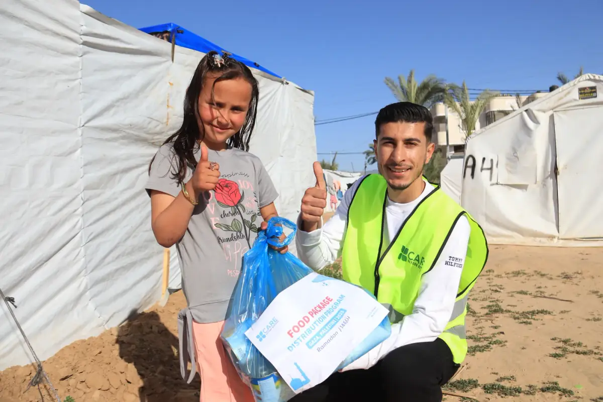 A smiling girl and a CBB volunteer give a thumbs up with her food package at a Gaza camp