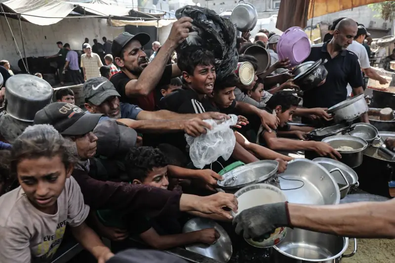 Children and adults reaching with empty pots at a meal distribution point in Gaza