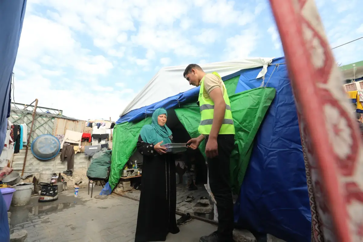 A CBB volunteer hands a hot-meal tray to a woman outside her tent in a Gaza displacement camp