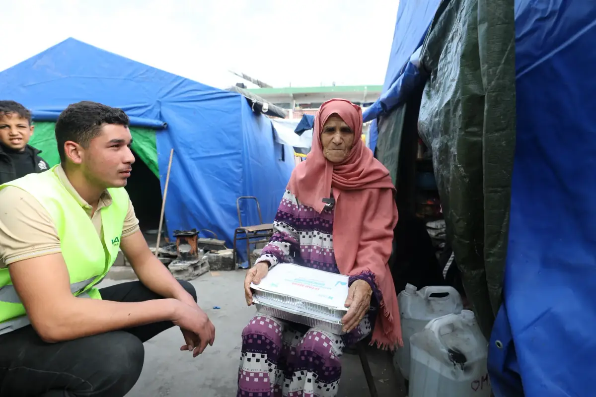 A CBB volunteer kneels beside an elderly woman holding a CBB iftar meal outside her tent in Gaza