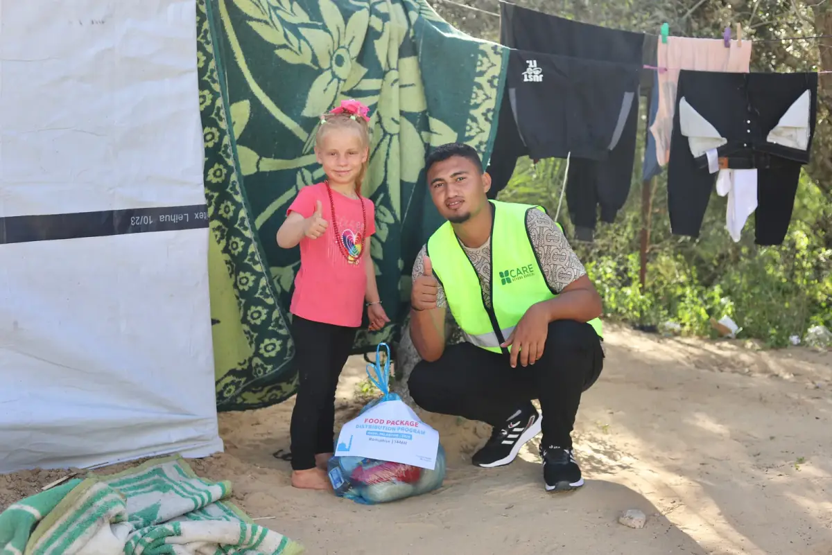A CBB volunteer with a young girl giving a thumbs up beside a food package outside her tent in Gaza