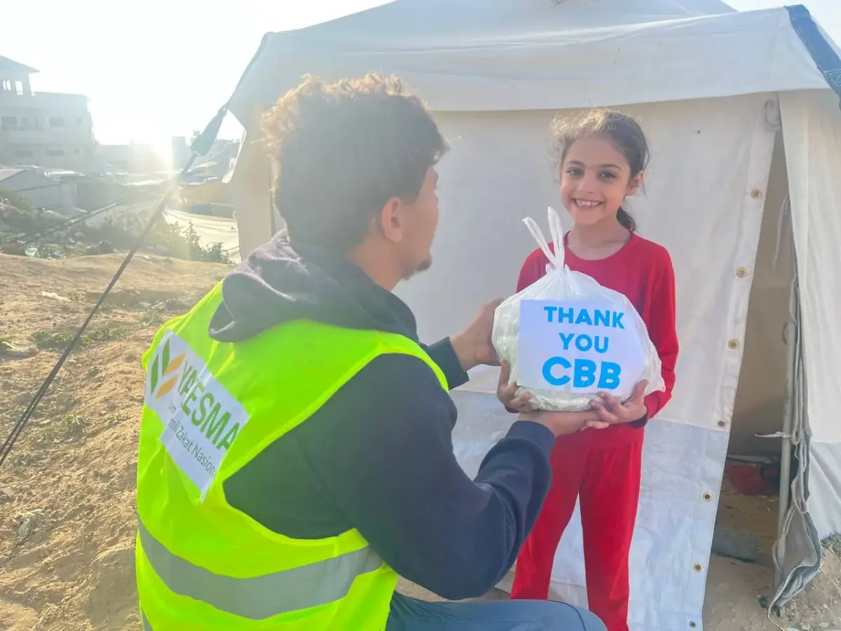 A CBB volunteer hands a "Thank you CBB" food package to a smiling girl outside her tent in Gaza